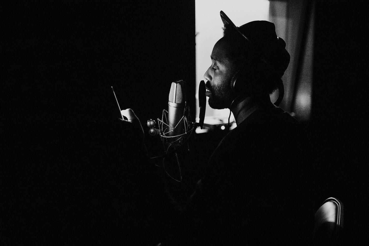 Black and white photo of a man recording vocals in a studio, wearing headphones and a hat, speaking or singing into a professional microphone.