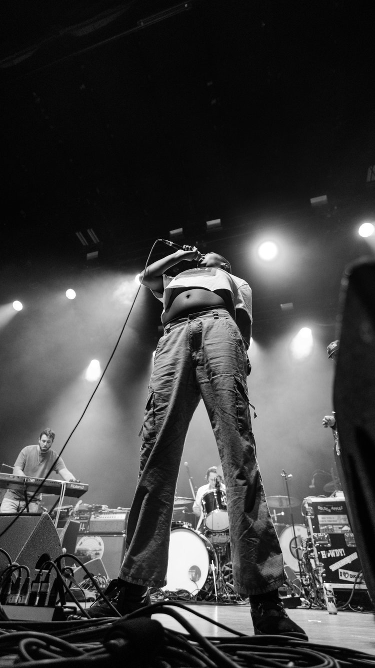 Black and white photo of a singer on stage holding a microphone above their head, with band members and musical equipment in the background.