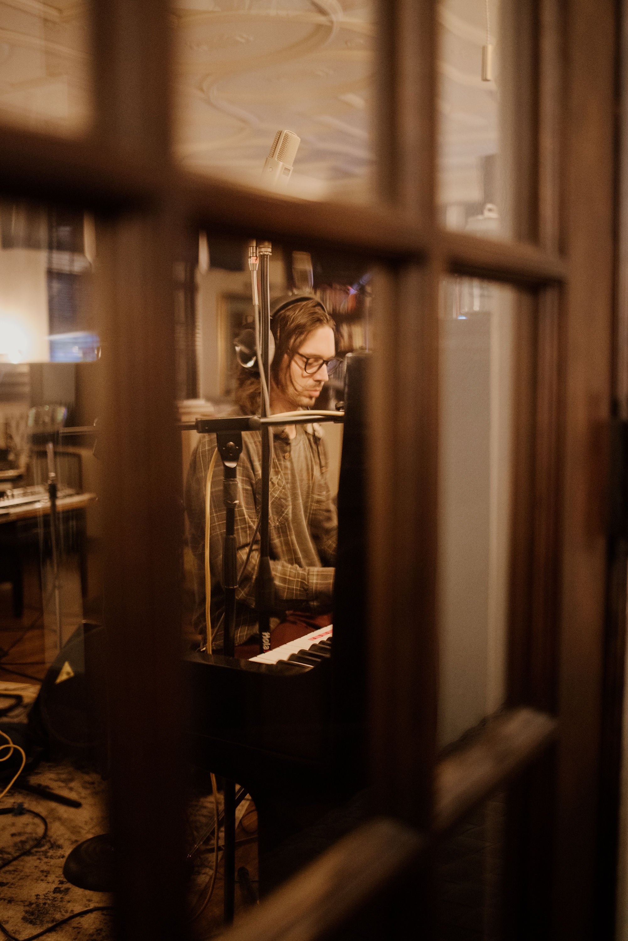 A man in glasses and a plaid shirt playing the piano in a recording studio, viewed through a wooden window frame.
