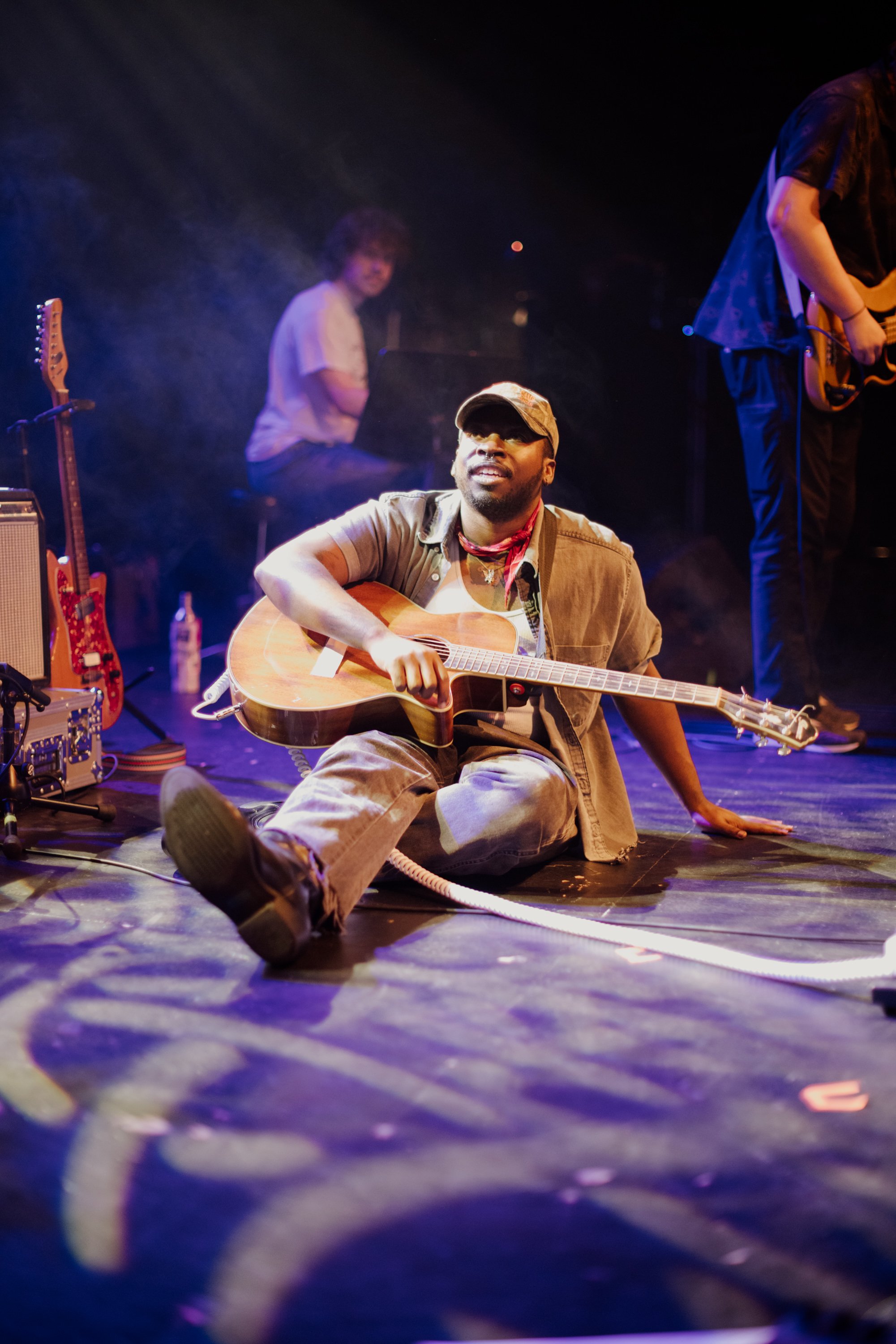 A musician sitting on stage with an acoustic guitar, smiling, with bandmates in the background. The stage is lit with colorful lights.
