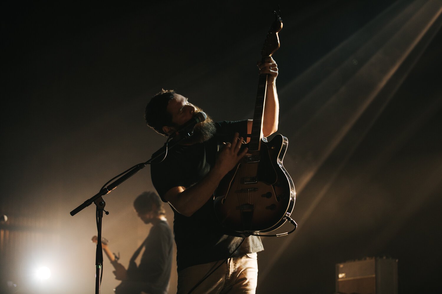 A man with a beard playing an electric guitar and singing into a microphone on stage, with another person playing guitar in the background, under concert lights.