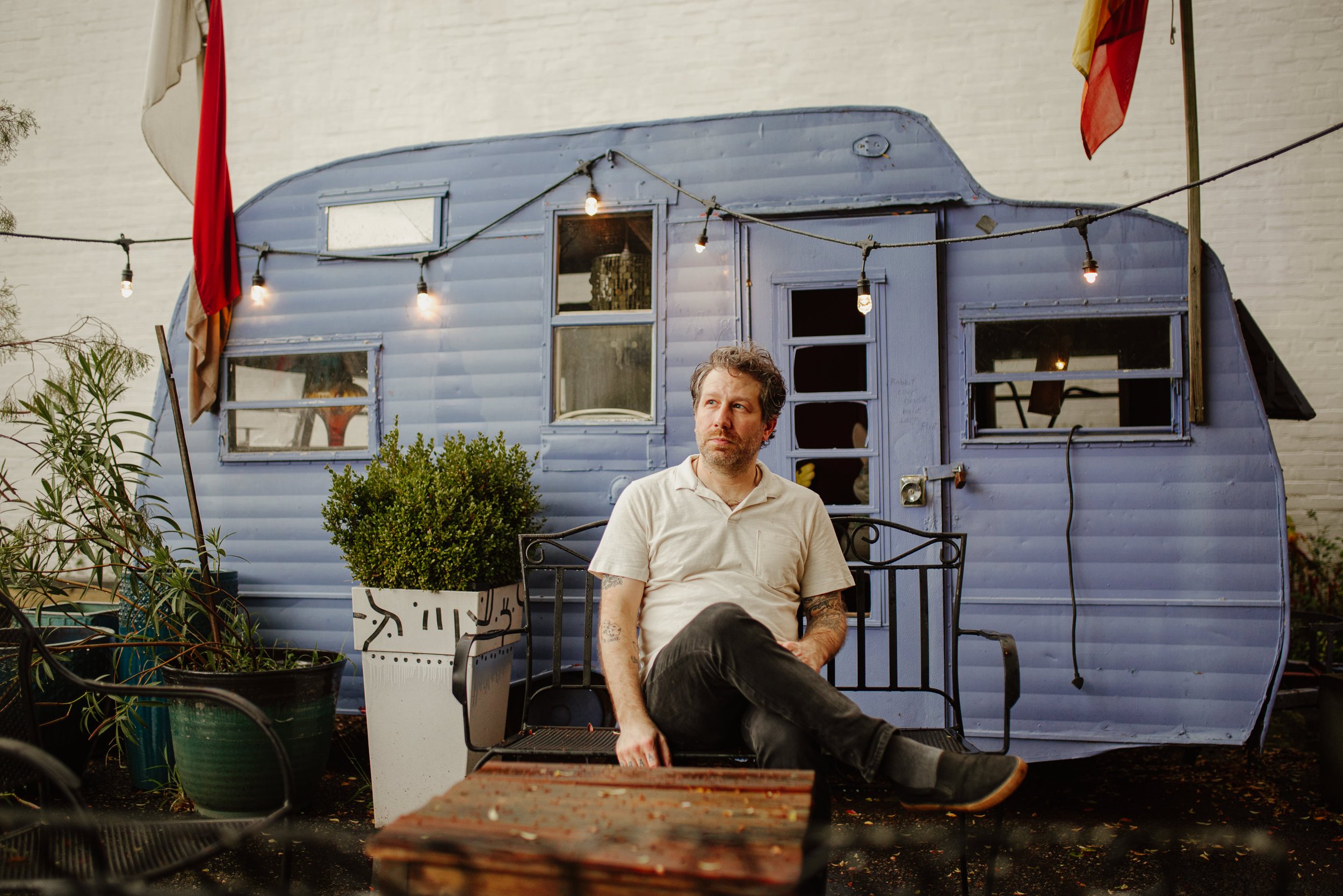Man sitting on a bench outside a vintage blue camper trailer, with string lights overhead and potted plants nearby.