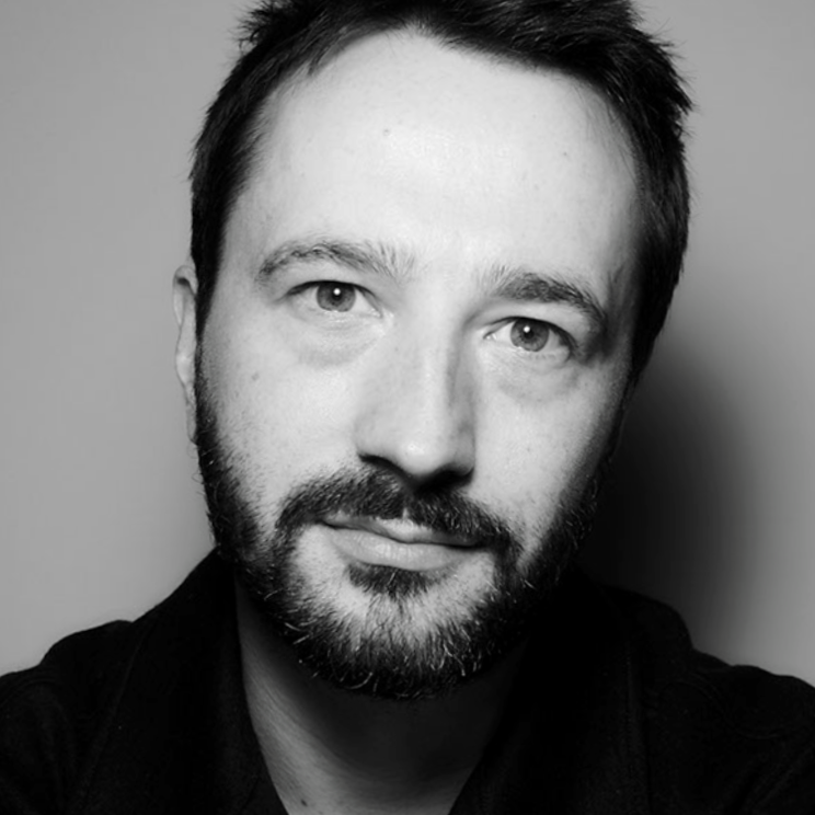 Black and white portrait of a man with dark hair and a beard, resting his chin on his hand, wearing a dark zip-up jacket, looking directly at the camera.