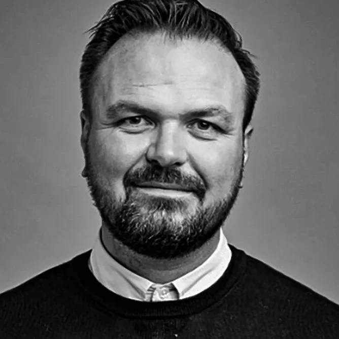 Black and white portrait of a man with short hair and a beard, wearing a collared shirt and a sweater, smiling slightly.