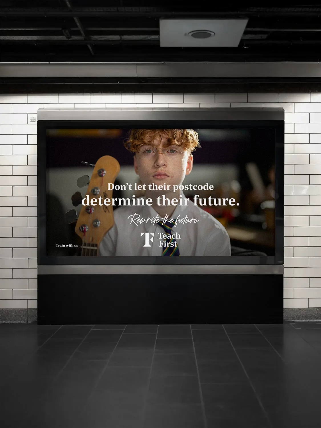 Digital billboard in a subway station displaying an advertisement for Teach First with a young person holding a guitar and text that reads, 'Don't let their postcode determine their future. Rewrite the future. Teach First.'