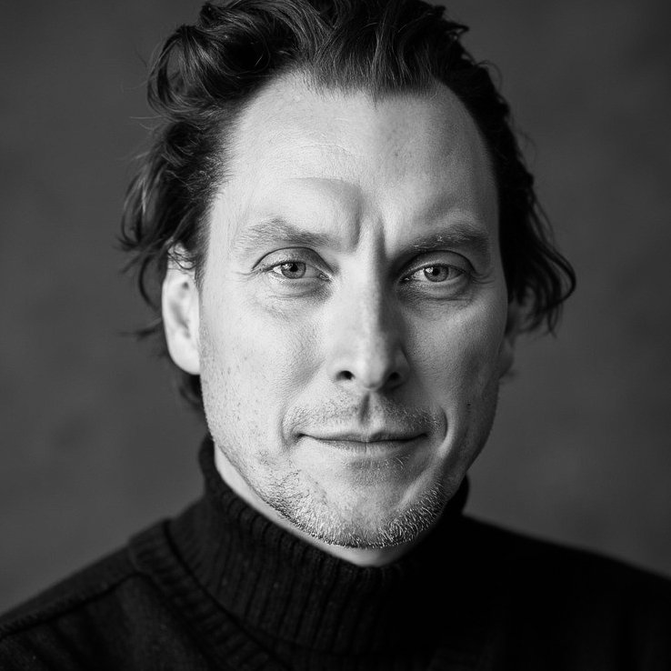 Black and white portrait of a man with short, wavy hair, wearing a turtleneck sweater, looking directly at the camera with a subtle smile.