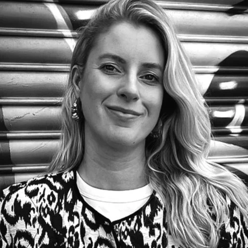 Black and white photo of a woman with long wavy hair, smiling, wearing earrings and a patterned top, standing in front of a metallic, corrugated background.