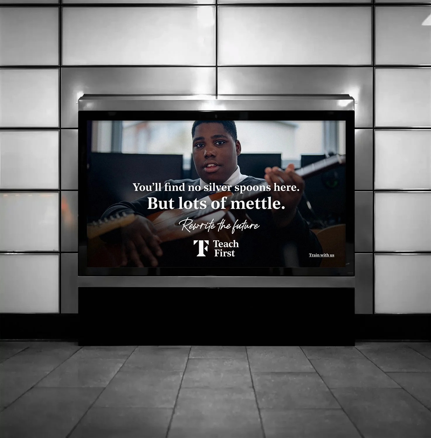 A digital display in an airport or train station showing an advertisement with a young person playing a guitar. The text on the ad reads, 'You'll find no silver spoons here. But lots of mettle. Rewrite the future. Teach First. Train with us.' The display is framed by metallic panels and is mounted on a tiled wall, with the floor visible in the foreground.