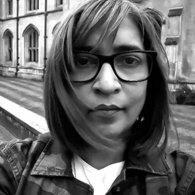 A woman with shoulder-length hair and glasses taking a selfie outdoors with a historic building in the background.