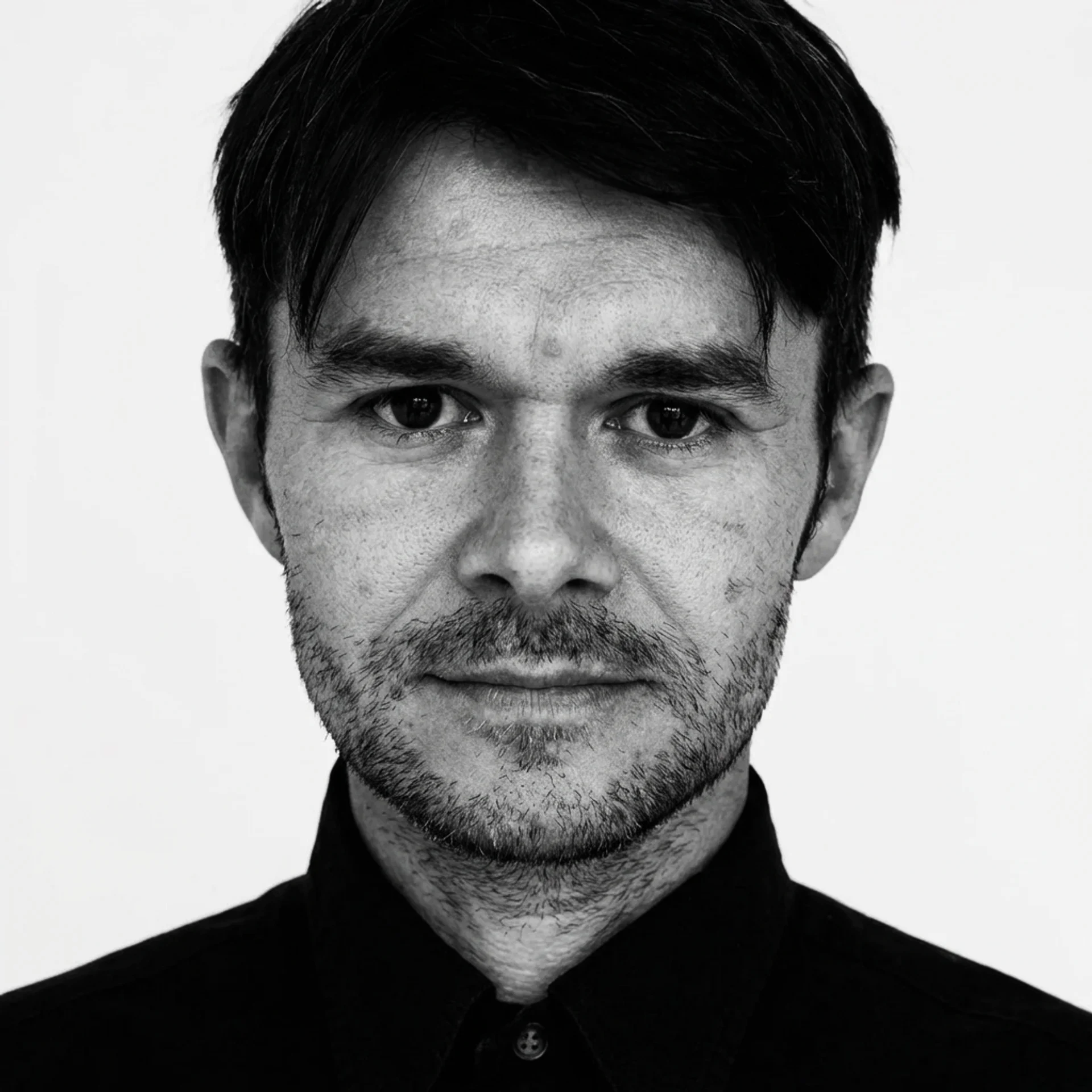 Black and white portrait of a man with dark hair and facial hair, wearing a collared shirt, looking at the camera against a plain background.