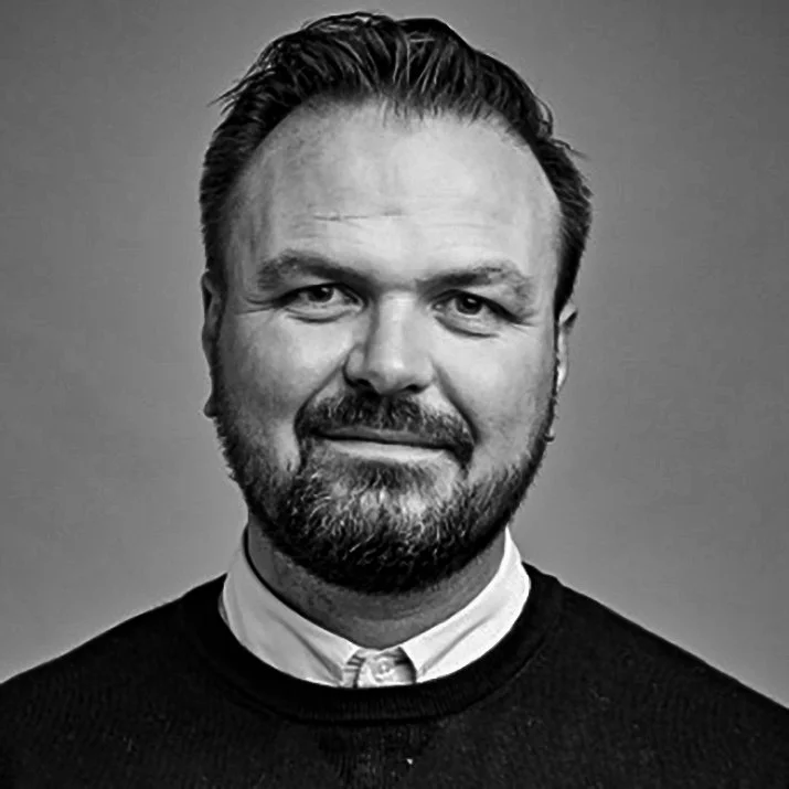 Black and white portrait of a man with short hair and a beard, wearing a collared shirt and a sweater, smiling slightly.