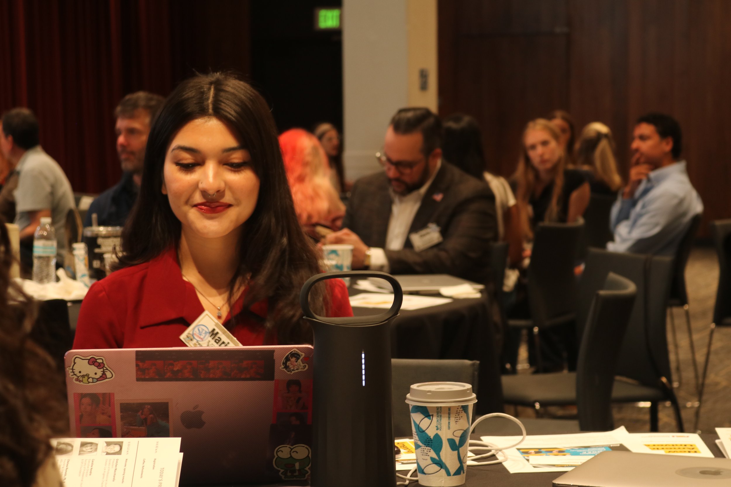 A woman works on her computer. Behind her a mix of men and women sit at round tables  taking notes and talking.