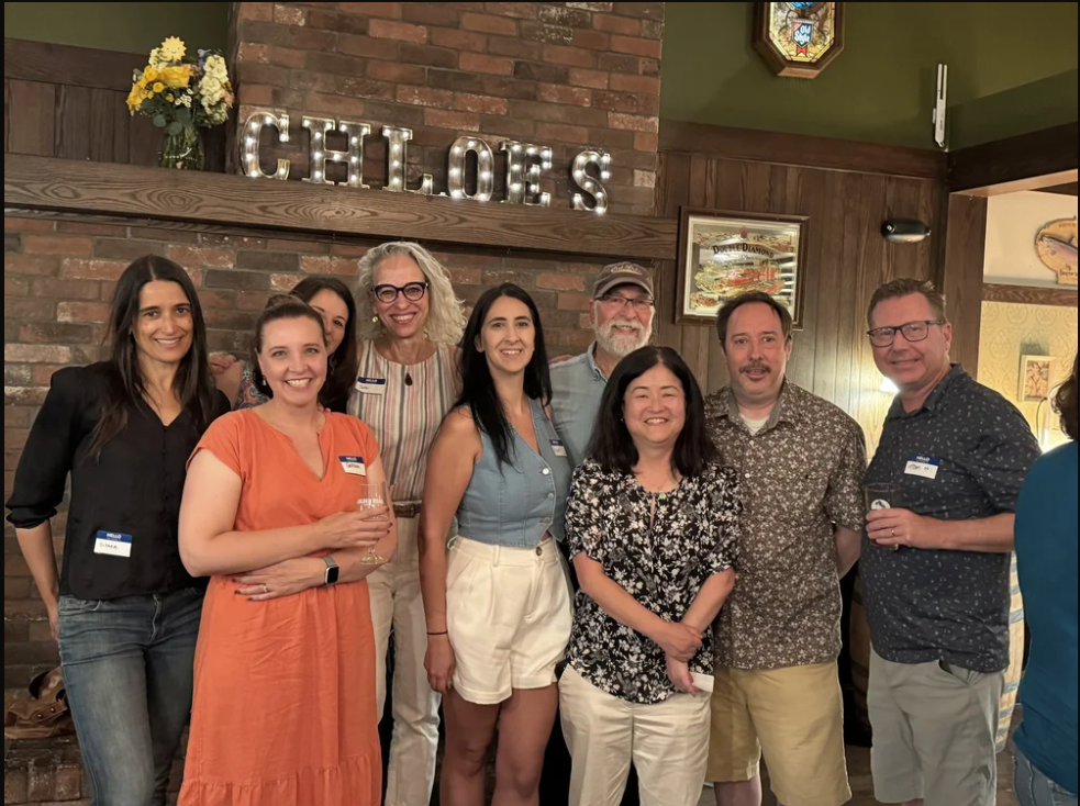 9 people stand in front of a brick wall and smile at the camera
