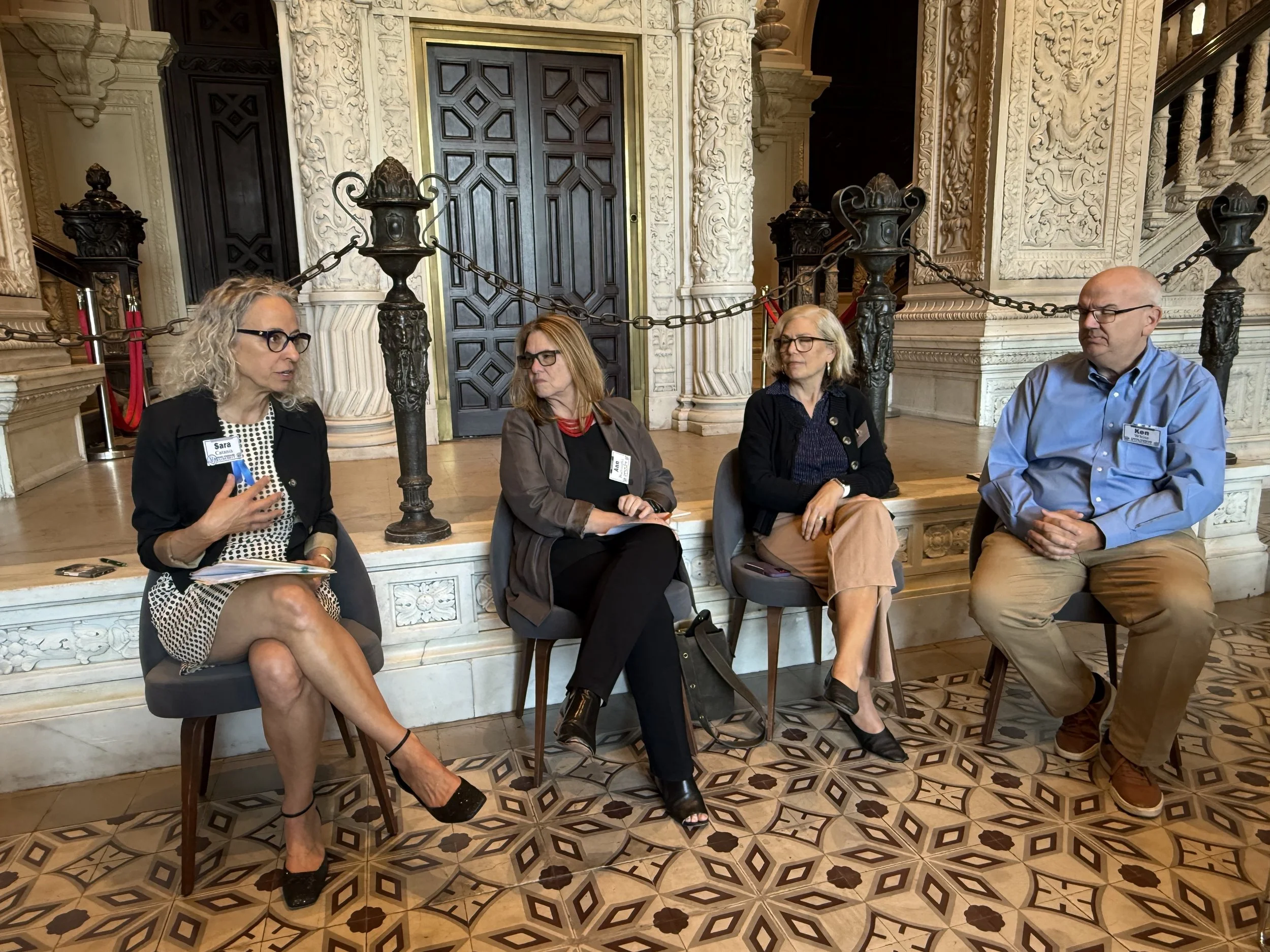 Three women and a man, all wearing glasses, sit in chairs and talk with a marble step and ornate marblework behind them.
