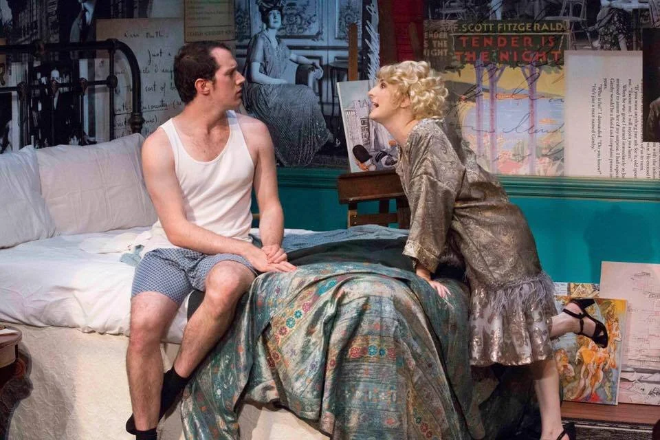 A man sitting on the edge of a bed looking surprised as a woman leans toward him, speaking and gesturing, in a bedroom with posters and artwork on the walls.