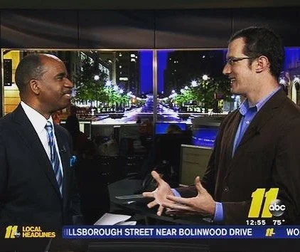 Two men engaged in conversation in a news studio, with a city street scene displayed on screens behind them.
