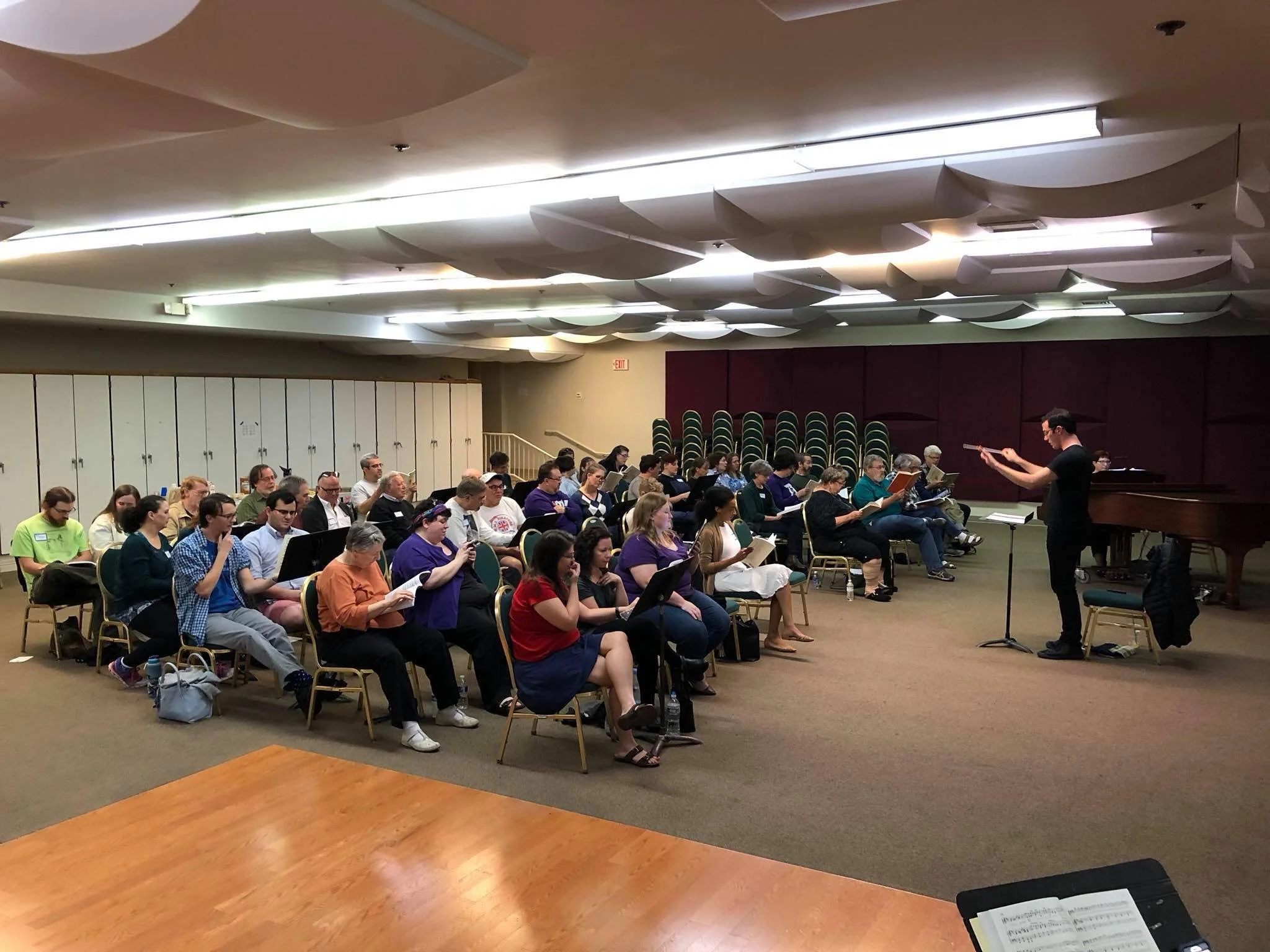 A conductor leads a rehearsal with a choir seated in rows inside a large room, with some chairs stacked against the back wall and a grand piano to the right.