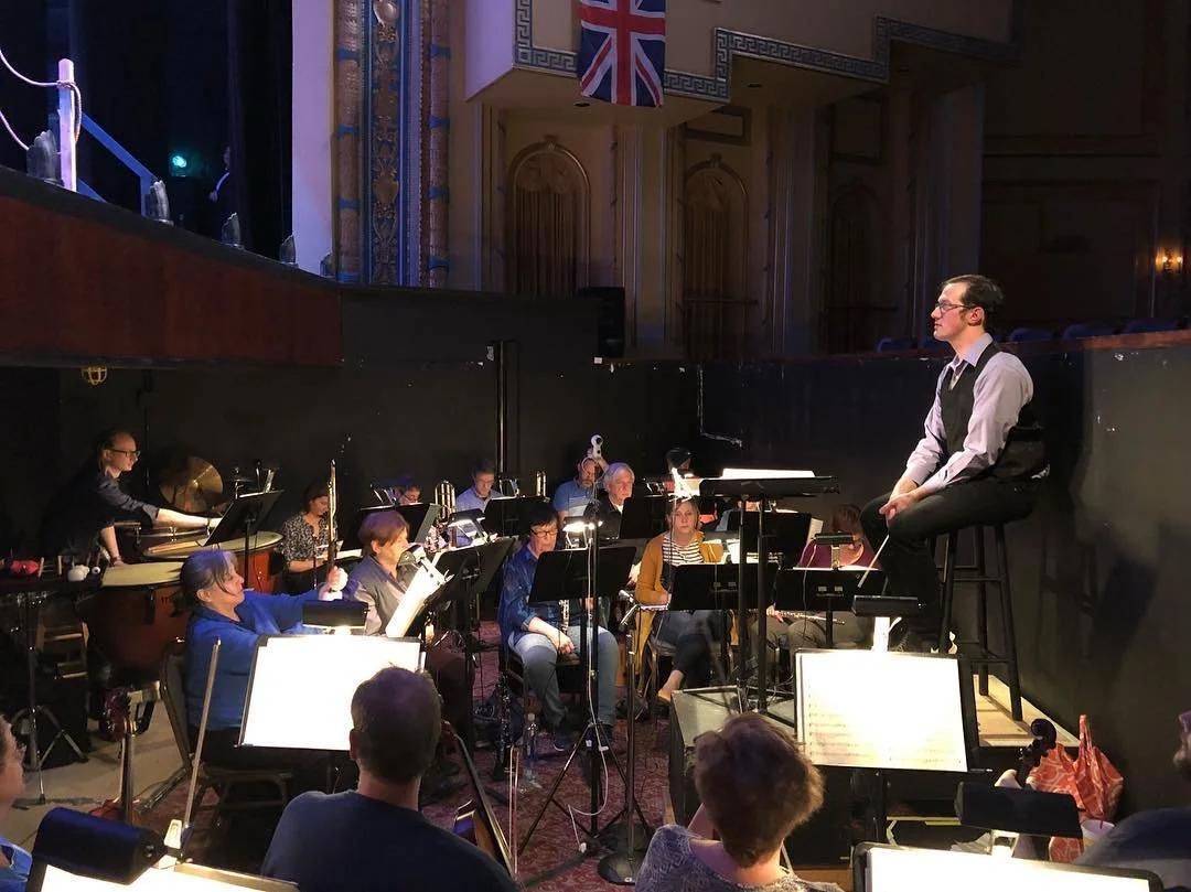 An orchestra performing on stage with musicians playing various instruments, and a conductor on a raised platform, in a concert hall with wooden paneling and a visible Union Jack flag.