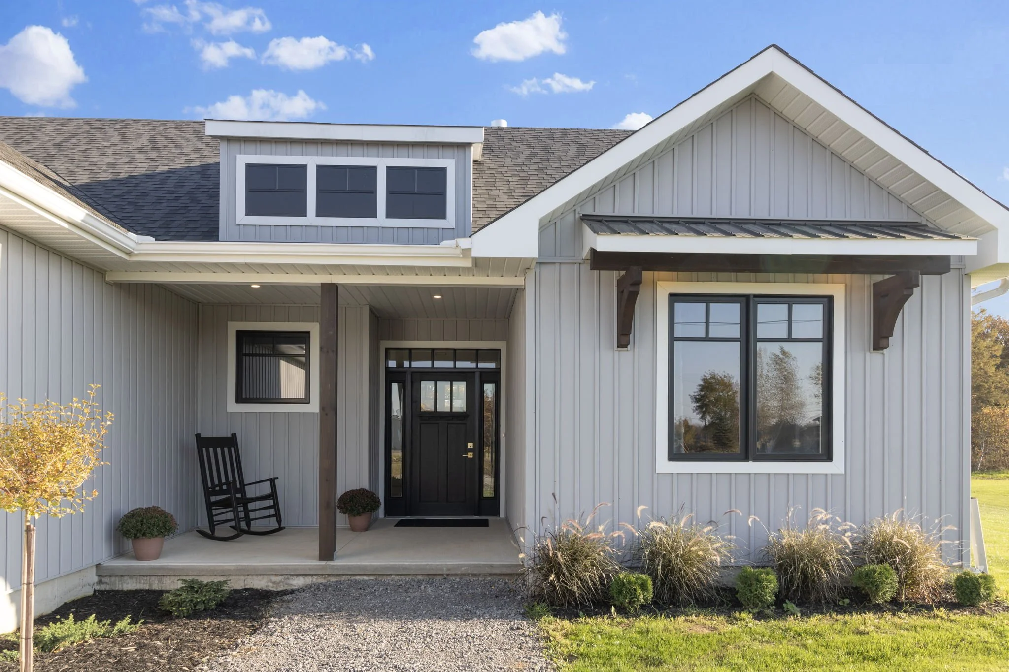 Front view of a modern farmhouse with vinyl siding, black front door, large front window, and small porch with a black rocking chair, surrounded by landscaped garden with bushes and plants, under a clear blue sky