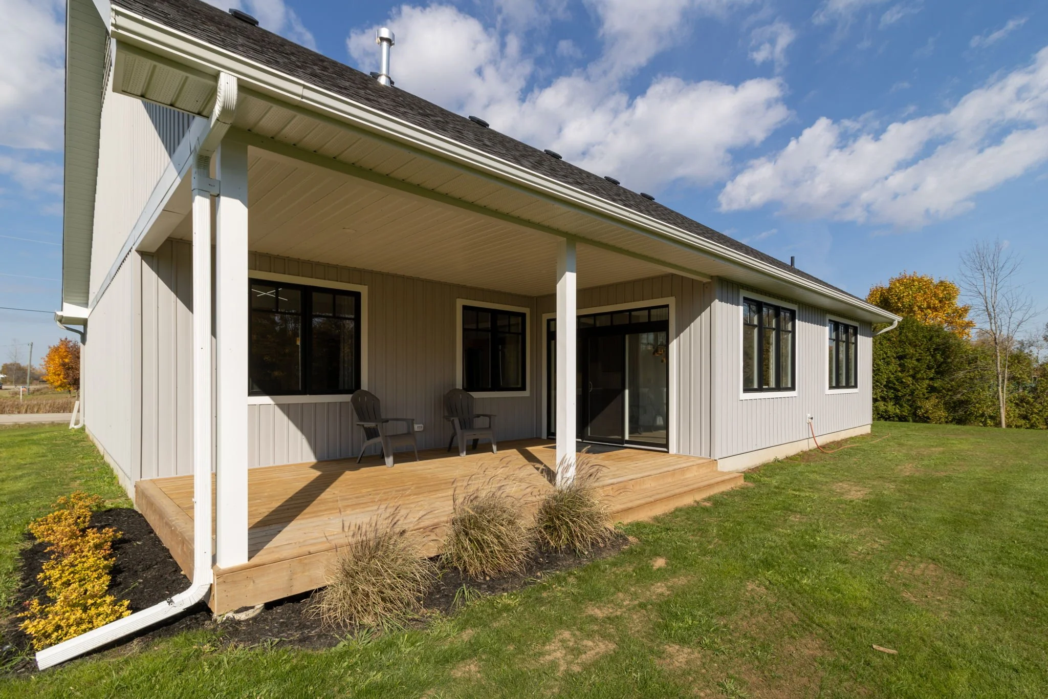 Back view of a modern house with a wooden deck, beige siding, black-framed windows, and a sliding glass door, surrounded by a lawn with a few small bushes, under a partly cloudy sky.