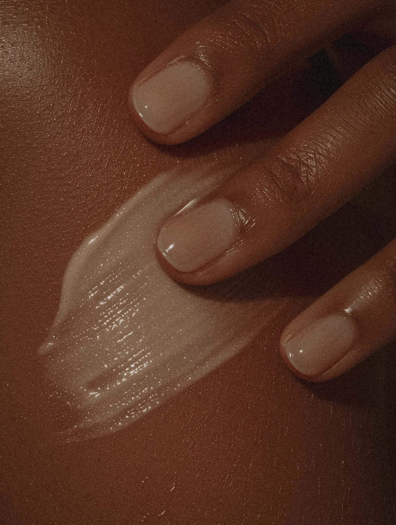 Close-up of three fingers with neatly manicured nails, with a shiny, light pink or nude nail polish, against a dark background.
