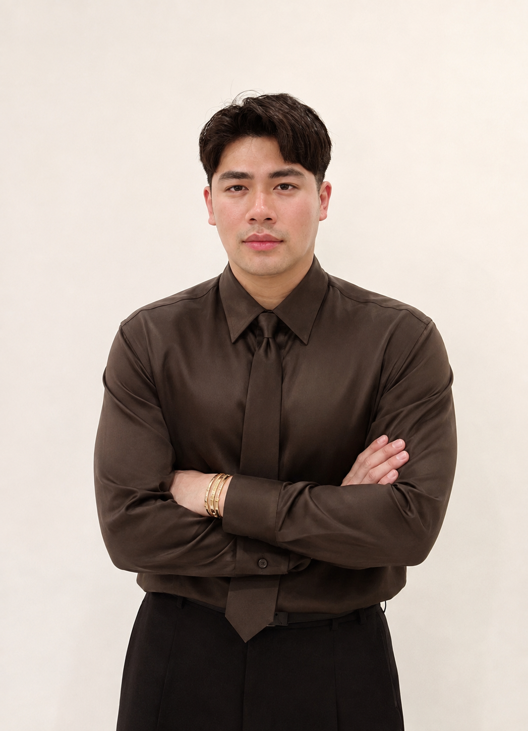 A young man with dark hair wearing a dark brown satin dress shirt and a matching tie, standing with his arms crossed against a plain light background.