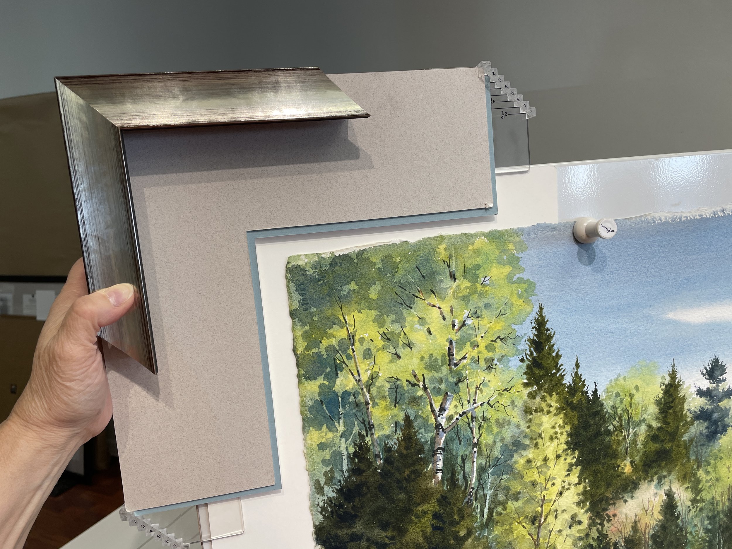 A hand holding a metallic picture frame corner with a beige mat board, positioned over a painting of a forest with trees and a blue sky.