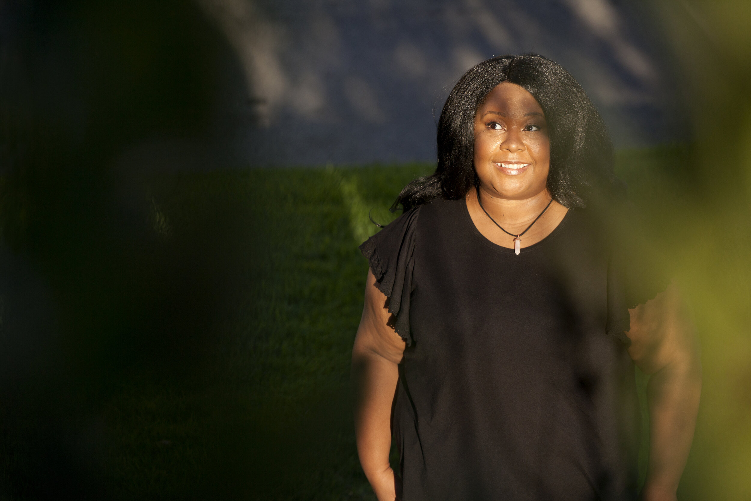 Brittany stands under a tree, with the shadow of a tree cascading across her face.  The sun peaks through the  leaves thought so she is still well-lit.  She is wearing a black tee, her hands to her side. She is facing forward but looking to the left.