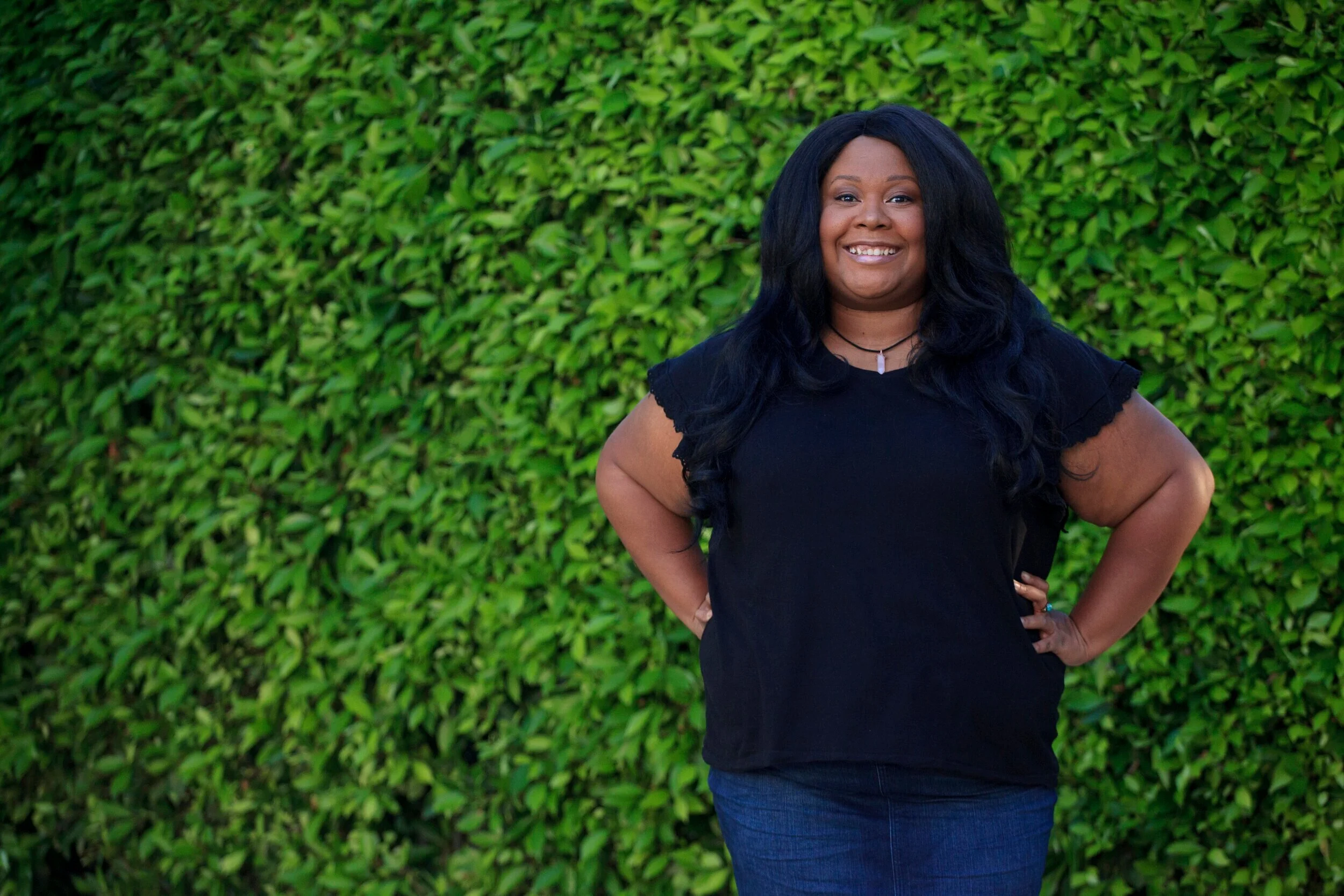 This is a photo of Brittany A Gash. A plus size, black woman with long black hair, a black tee shirt, stands with her hands on her hips. She is smiling.  A green hedge wall is in the background.