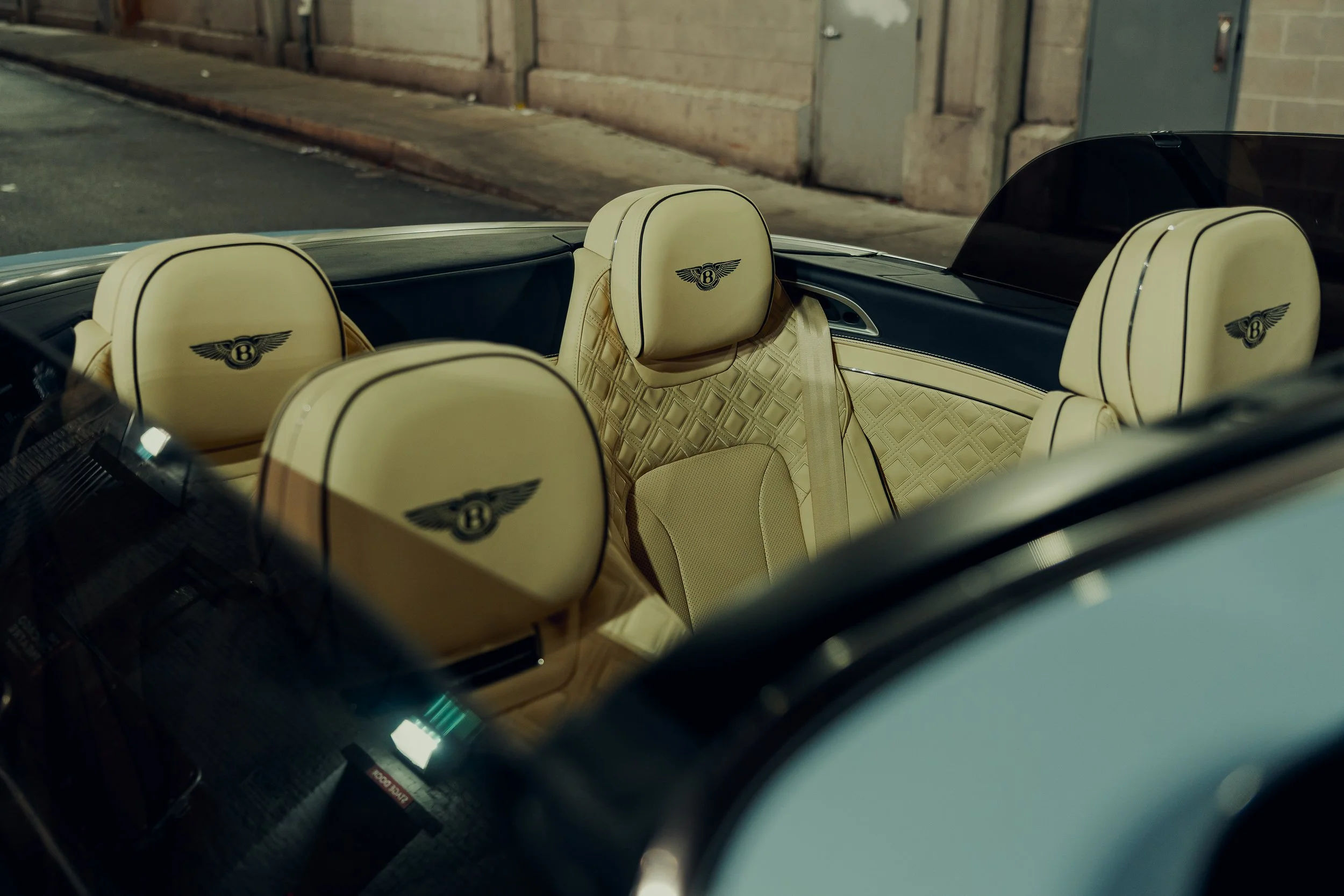 The interior of a luxury convertible car with cream-colored seats featuring the Bentley logo, parked on a street at night.