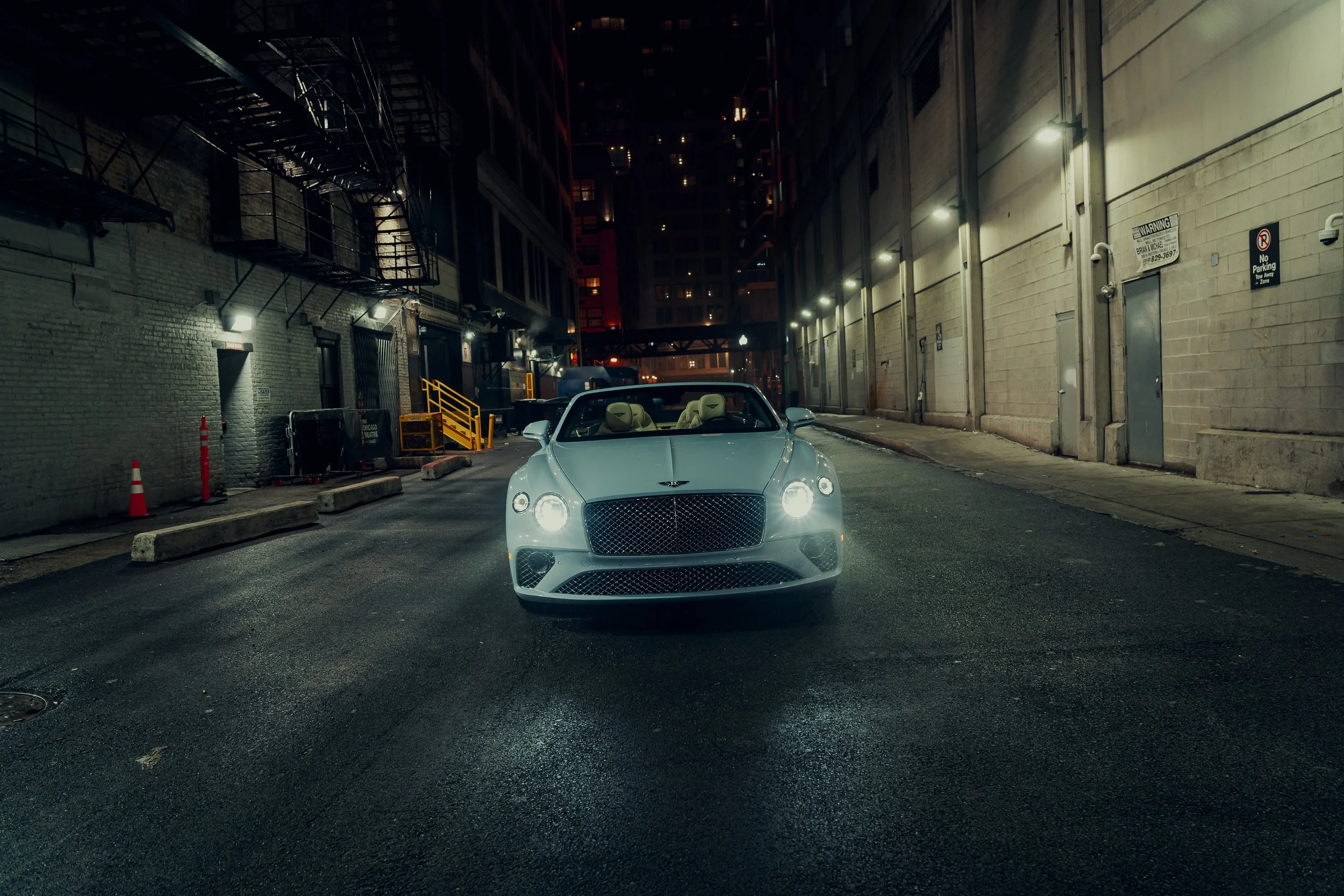A white convertible luxury car parked on a city street at night, with dim lighting, brick walls, fire escape stairs, and tall buildings in the background.