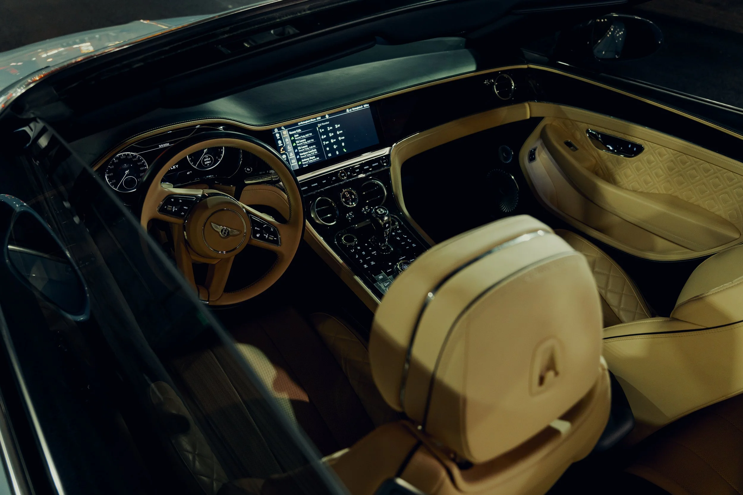 The interior of a luxury car featuring a beige leather dashboard, steering wheel with a Bentley emblem, large digital display screen, and beige leather seats with quilted pattern.