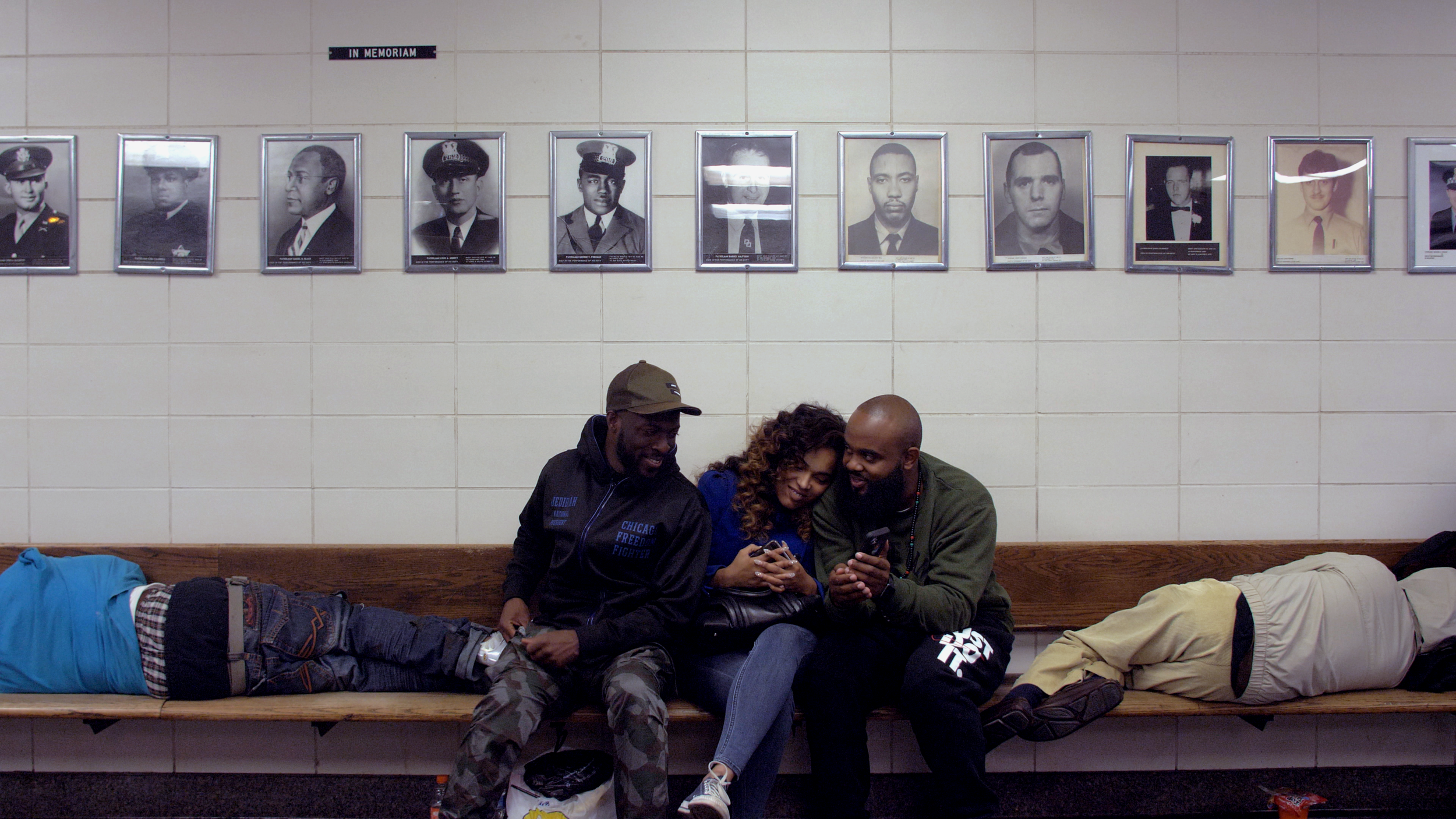 Three people sitting on a bench in a memorial lobby, with photographs of officers on the wall behind them, two people lying down on the bench and two people looking at a phone.