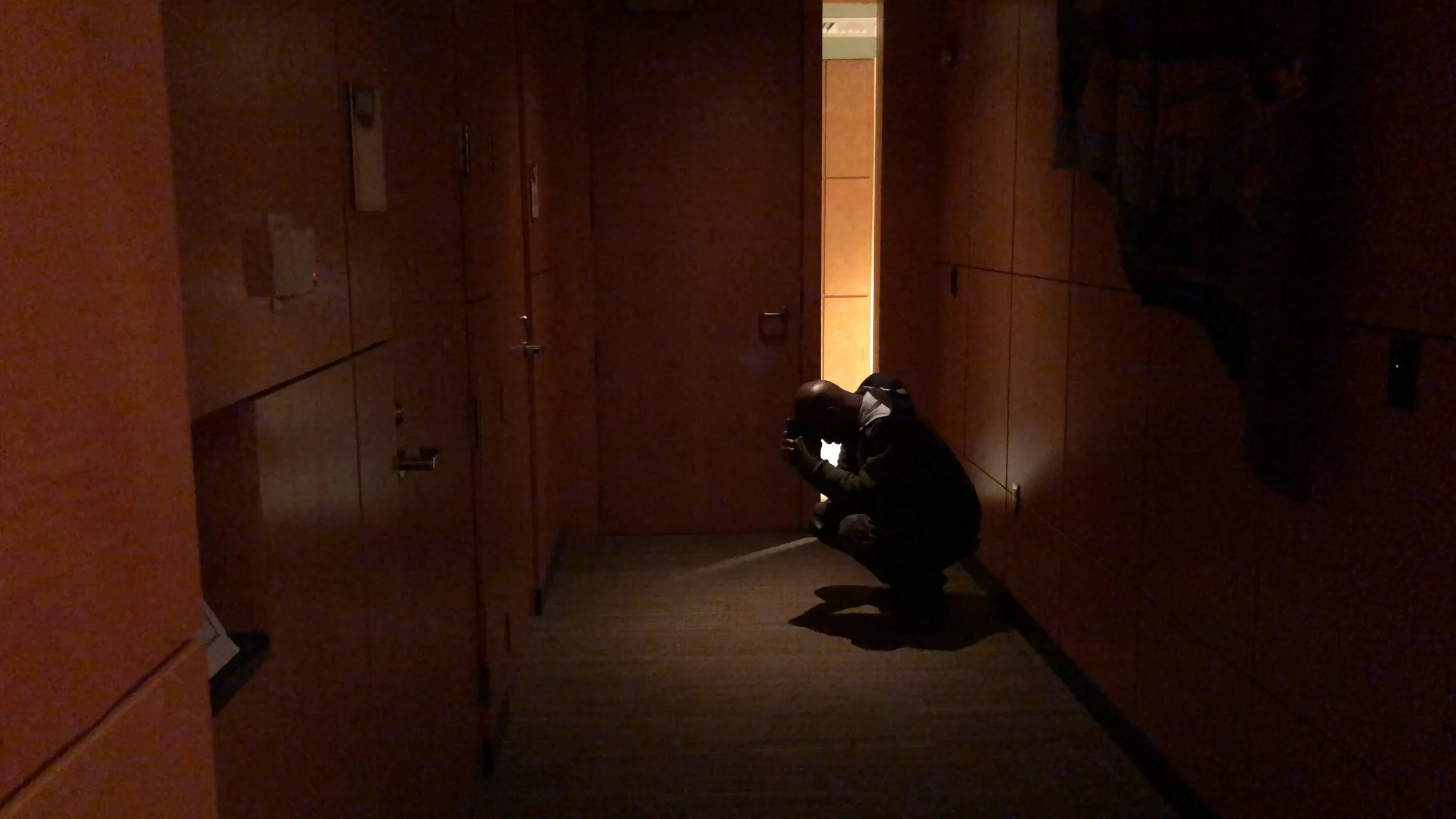 A man crouching in a dimly lit corridor, taking a photo or looking at something on his phone, with wooden cabinets on each side.