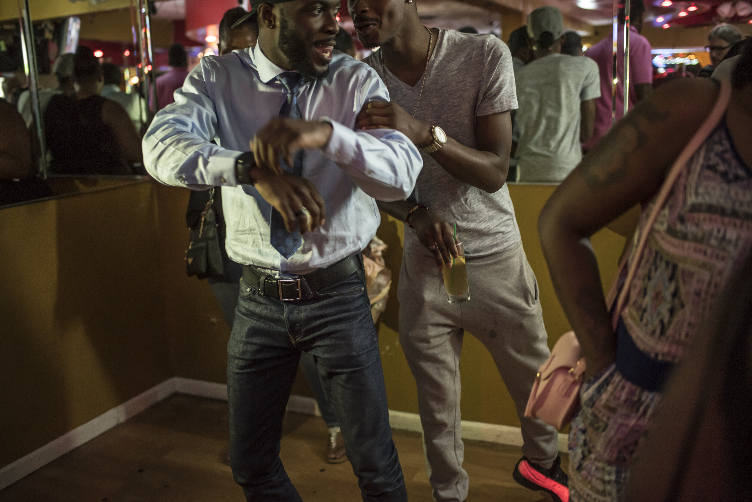 Three people dancing and socializing at a lively indoor party. One man in a dress shirt and tie is dancing with another man dressed casually, holding a drink. A woman wearing a patterned dress and carrying a pink purse is partially visible on the rig
