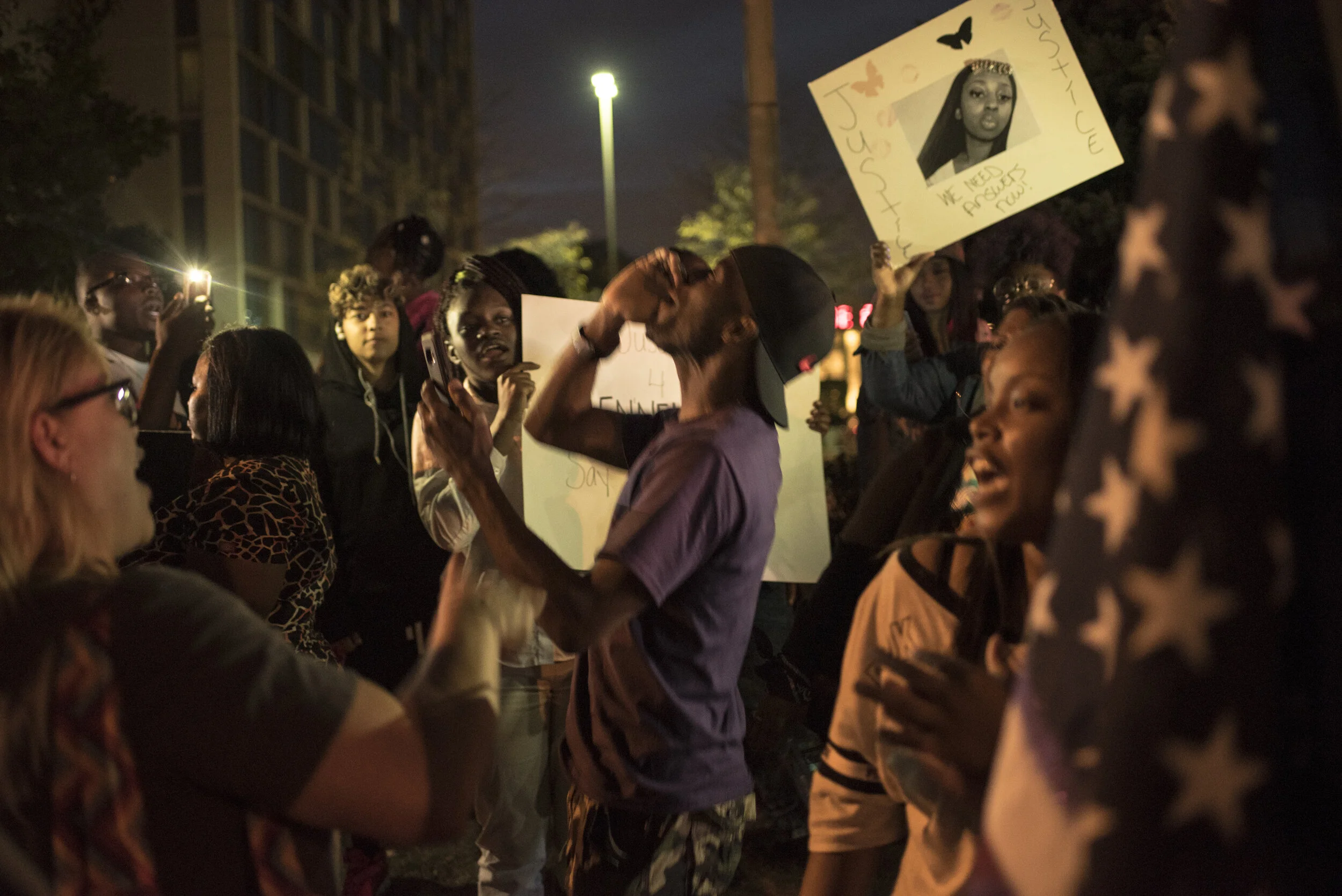 A diverse group of people gathered outdoors at night, some holding signs related to social justice, while engaging in conversations and taking photos. The scene appears to be a peaceful protest or rally.