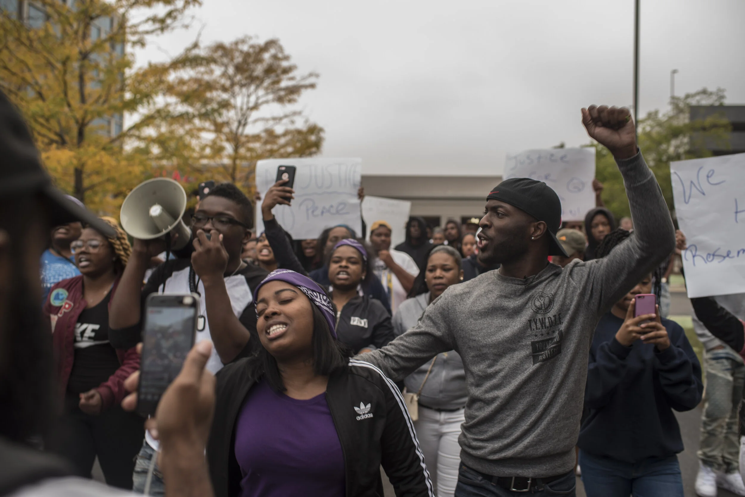 A group of people protesting, some with raised fists and others holding signs that read "Justice" and "Peace". The crowd appears to be engaged and passionate, with some individuals recording or taking photos of the event.