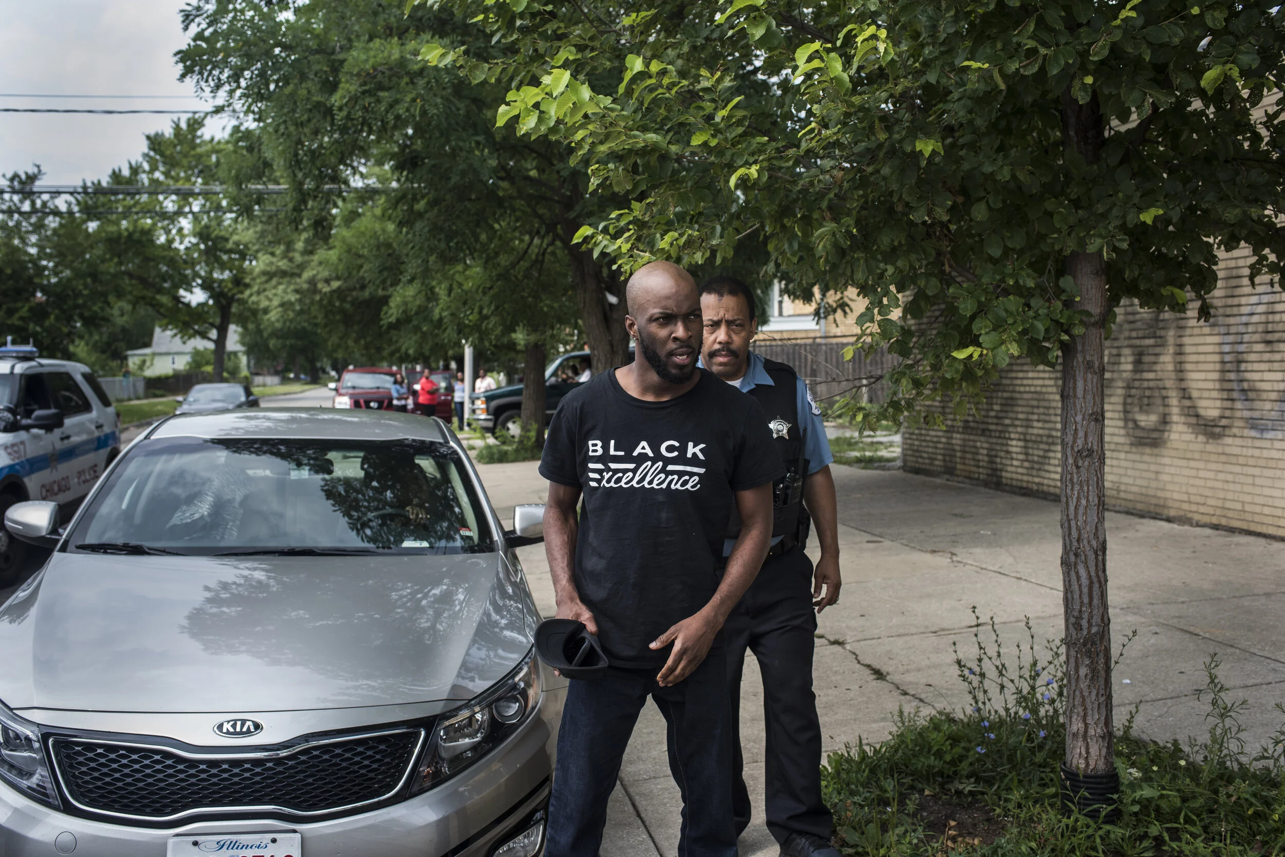 A man with a shaved head and a beard, wearing a black T-shirt with the words 'Black Excellence,' stands on a sidewalk next to a silver Kia car, with a police officer behind him. The scene takes place in a neighborhood with trees, parked cars, and a b