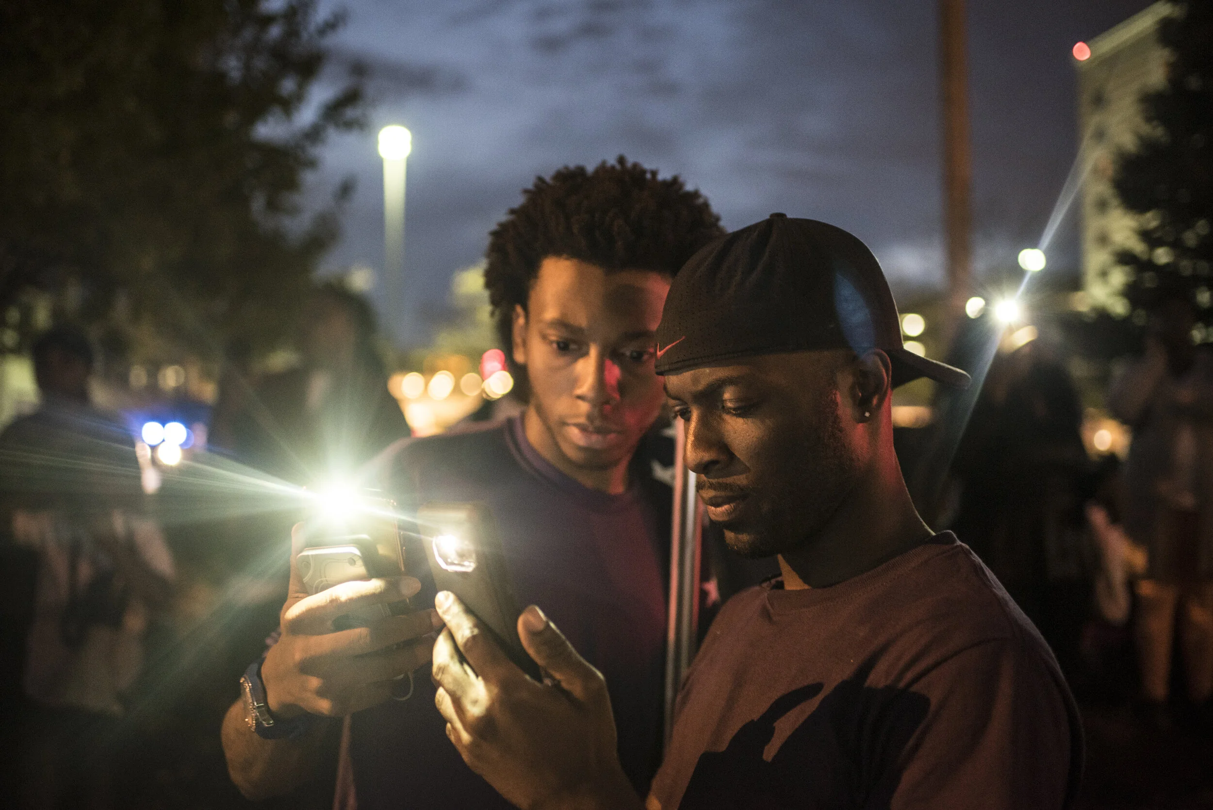 Two men look at a phone screen during a nighttime outdoor gathering, with one man wearing a backwards cap and the other with curly hair. City lights and other people are visible in the background.