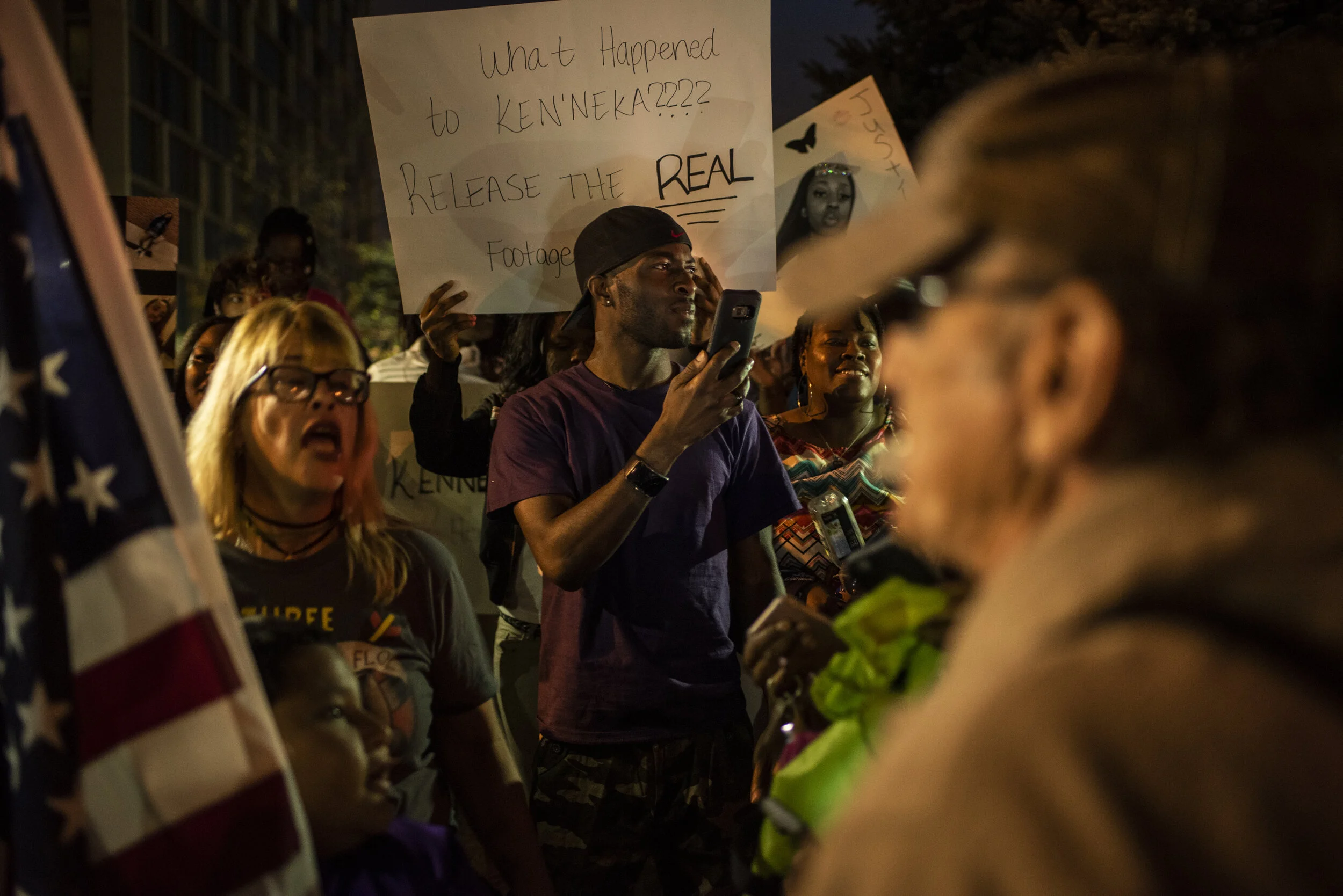 Crowd of protesters gathered at night, with one person holding a sign that reads 'What Happened to KENNEKA??? RELEASE THE REAL Footage,' and another person speaking into a microphone.
