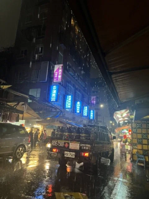 Rainy street scene in a city at night with illuminated signs, a truck, and people with umbrellas.
