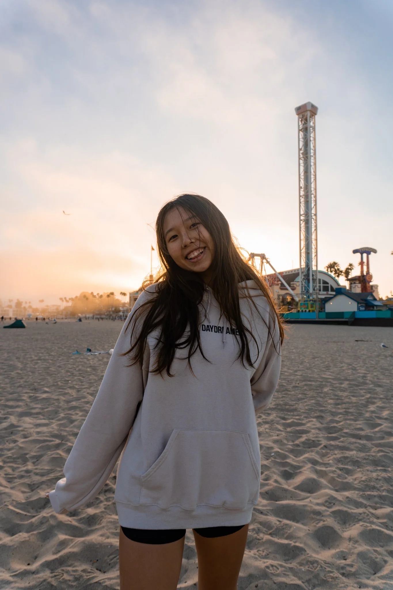 Smiling person in a beige hoodie on a sandy beach during sunset with amusement park rides in the background.