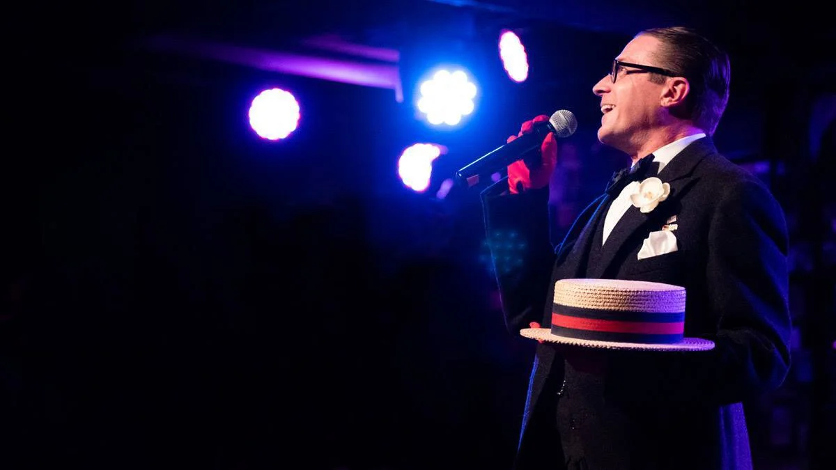 Dapper white middle-aged man in profile holding microphone in red-gloved hand and straw boater hat in the other. Stage lights visible in background.
