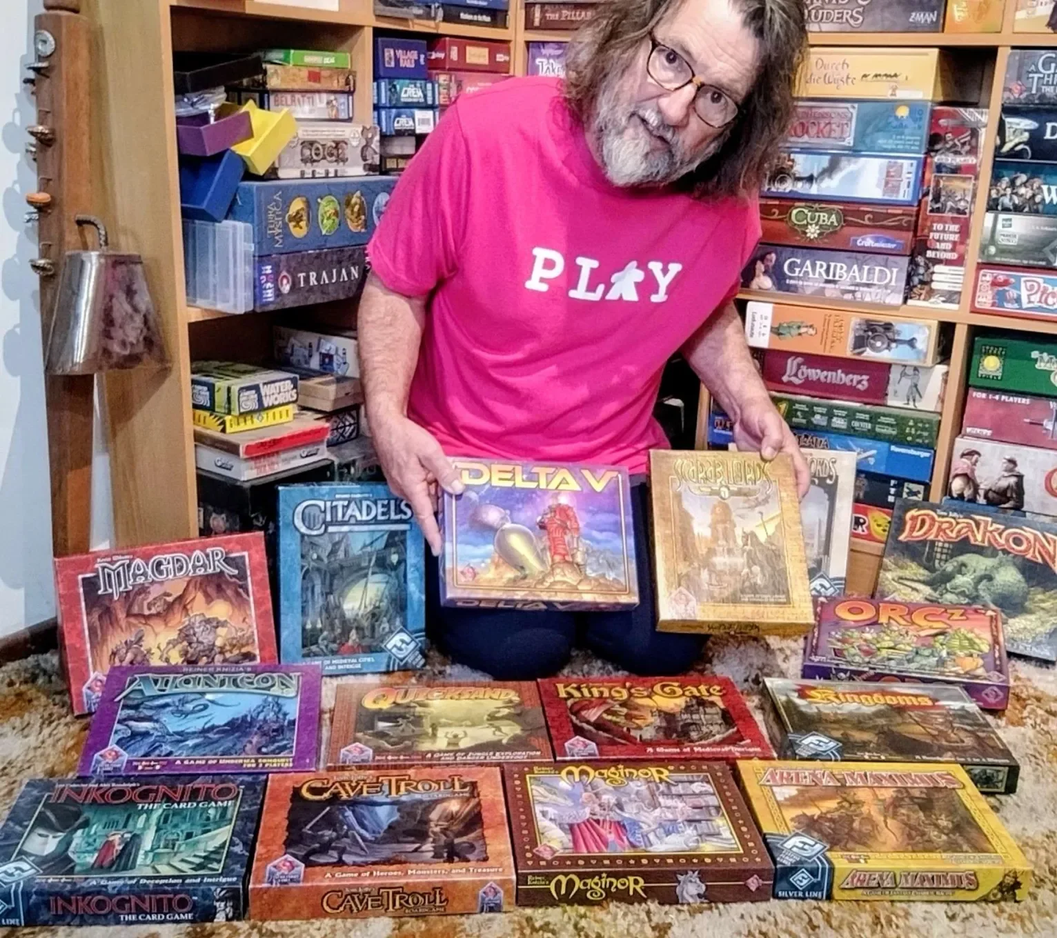 Elderly white dude in glasses and fuschia T-shirt reading PLAY, shows off several square vintage board game boxes from Fantasy Flight Games' Silver Series
