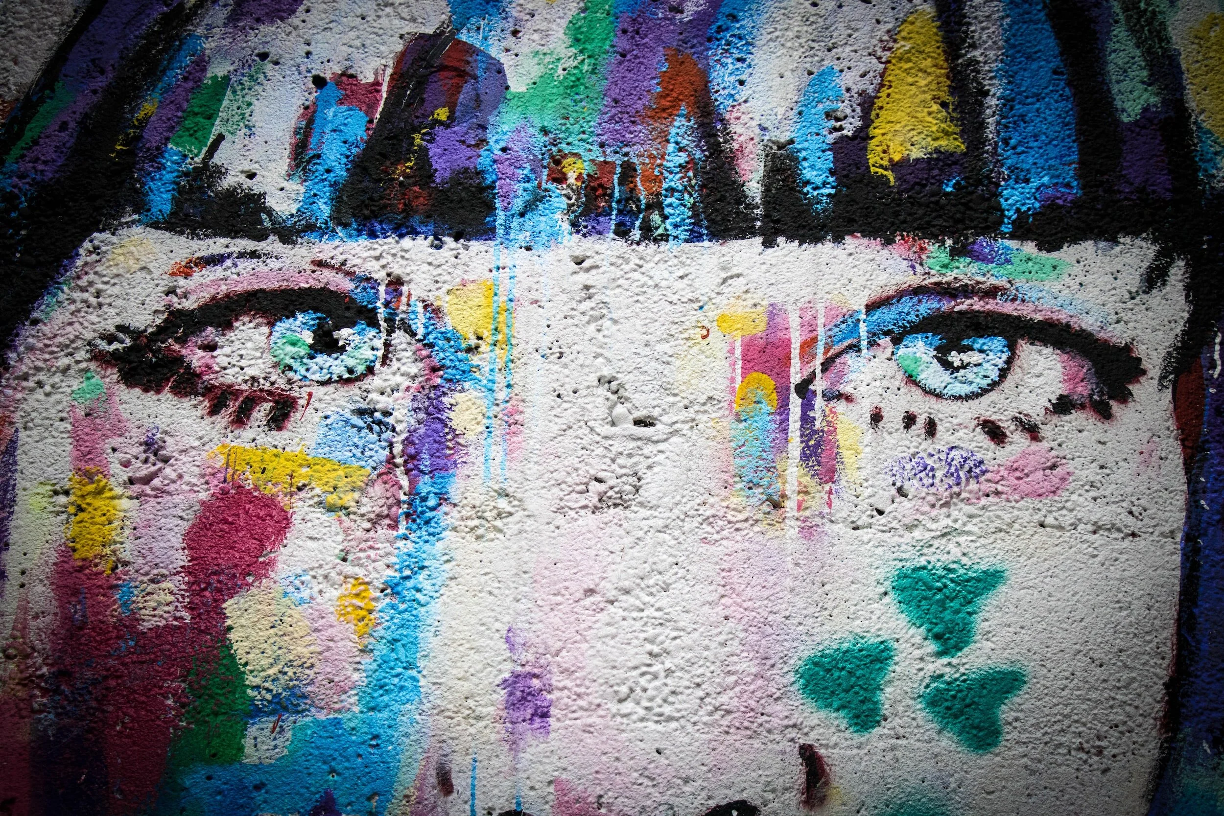 Image description shows a spray painting on a rough, white wall, of bright colors including aqua blue, pink, and yellow, and black. The paining is of a human face from the nose to the hairline. The eyes are gazing straight forward.