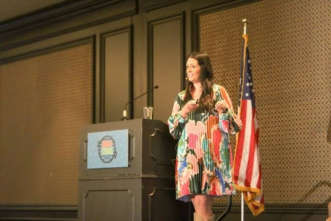 A woman standing on a stage during a conference, holding a microphone. Behind her are four vertical screens with the words 'Brooke Taylor' and logos, and a purple curtain backdrop. Audience members are visible in the foreground.
