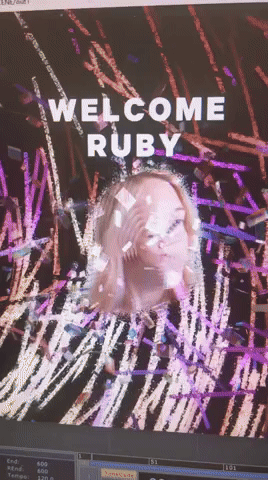 Screen displaying a message that says "WELCOME RUBY" with a digital rainbow-colored background and an overlayed image of a woman making a peace sign.