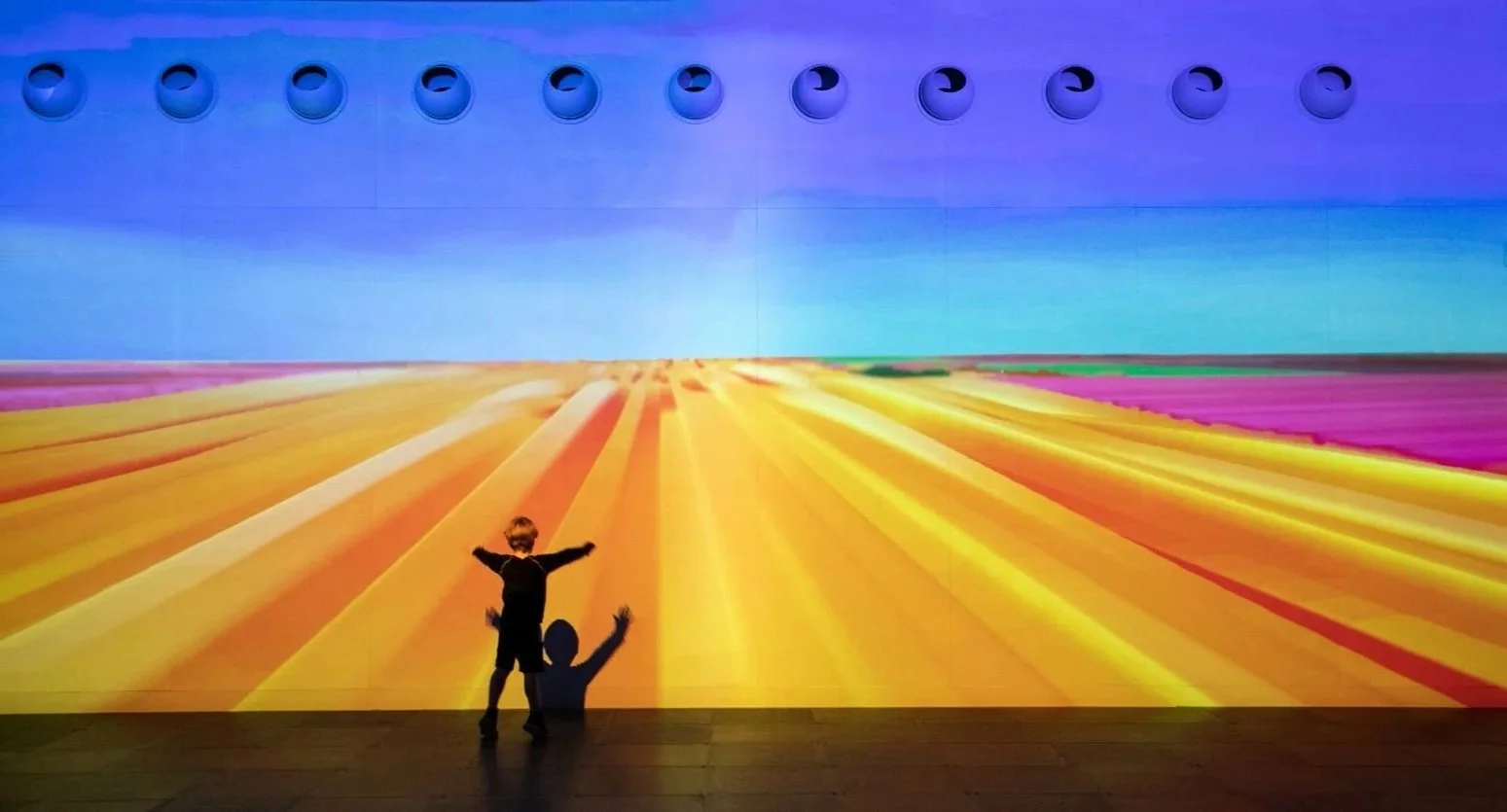 Two children standing in front of a large digital art installation depicting colorful fields and a blue sky.