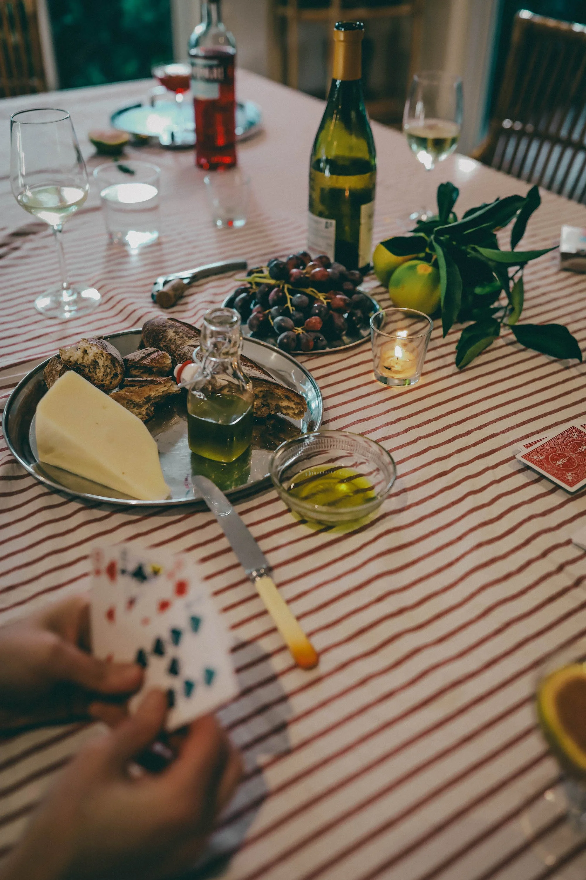 Bread, cheese, olive oil, wine and grapes on a striped tablecloth by candlelight, with cards being dealt