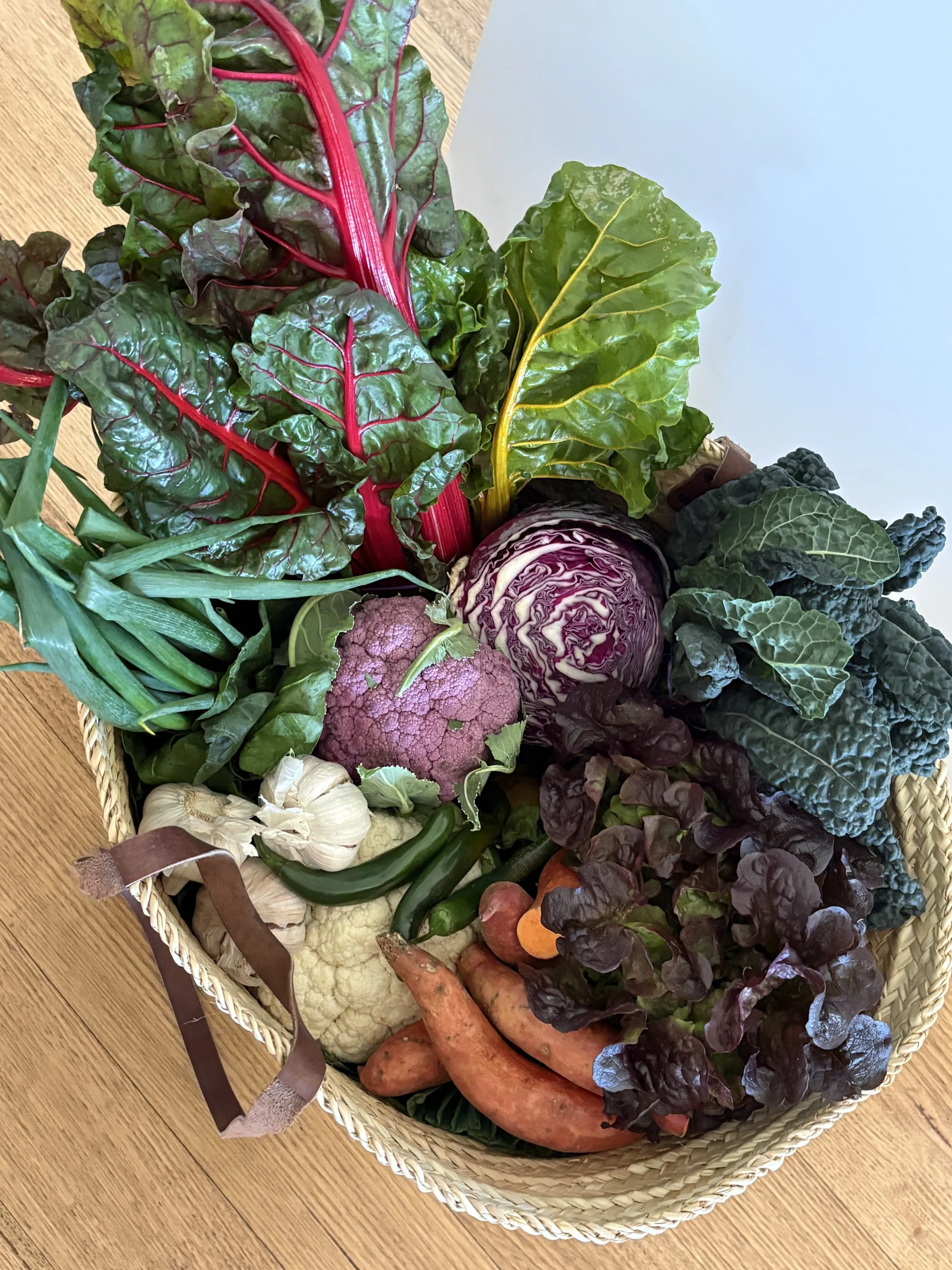 Overhead view of a woven basket of vegetables from Churchill Island Market — rainbow chard, purple cauliflower, red cabbage, kale and sweet potatoes